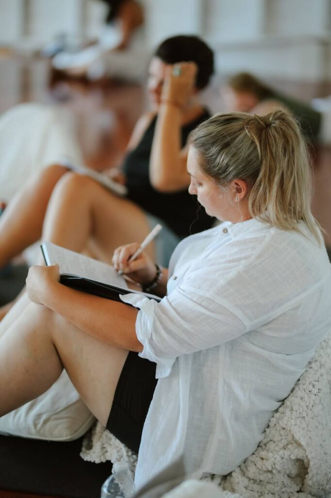 woman in a white shirt and black shorts, blonde hair in a ponytail, sitting on the floor against a cushion.  she is writing in a journal that is resting on her knees. 