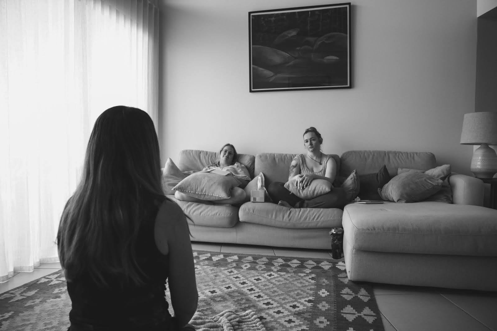 black and white image of a woman, sitting close to the camera but facing away, looking towards to women sitting across from here who are looking calm and serious.